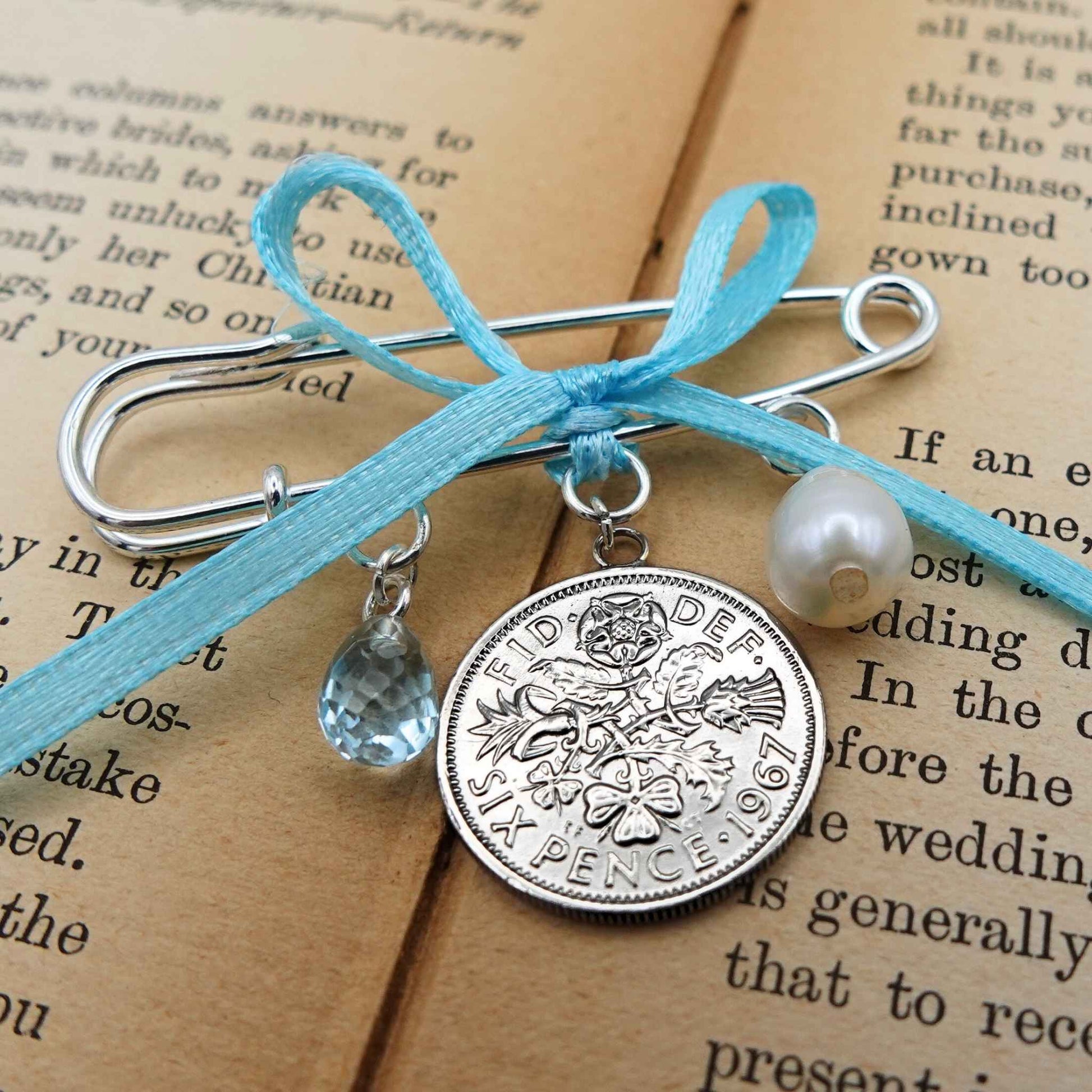 Bridal brooch with British sixpence coin charm, white freshwater pearl, and blue topaz with a blue ribbon, placed on a book page.