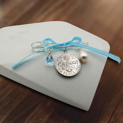 White freshwater pearl, blue topaz teardrop, genuine Queen Elizabeth 2nd sixpence coin with baby blue ribbon all suspended from a silver plated safety pin. Bridal safety pin on tile with wooden background.
