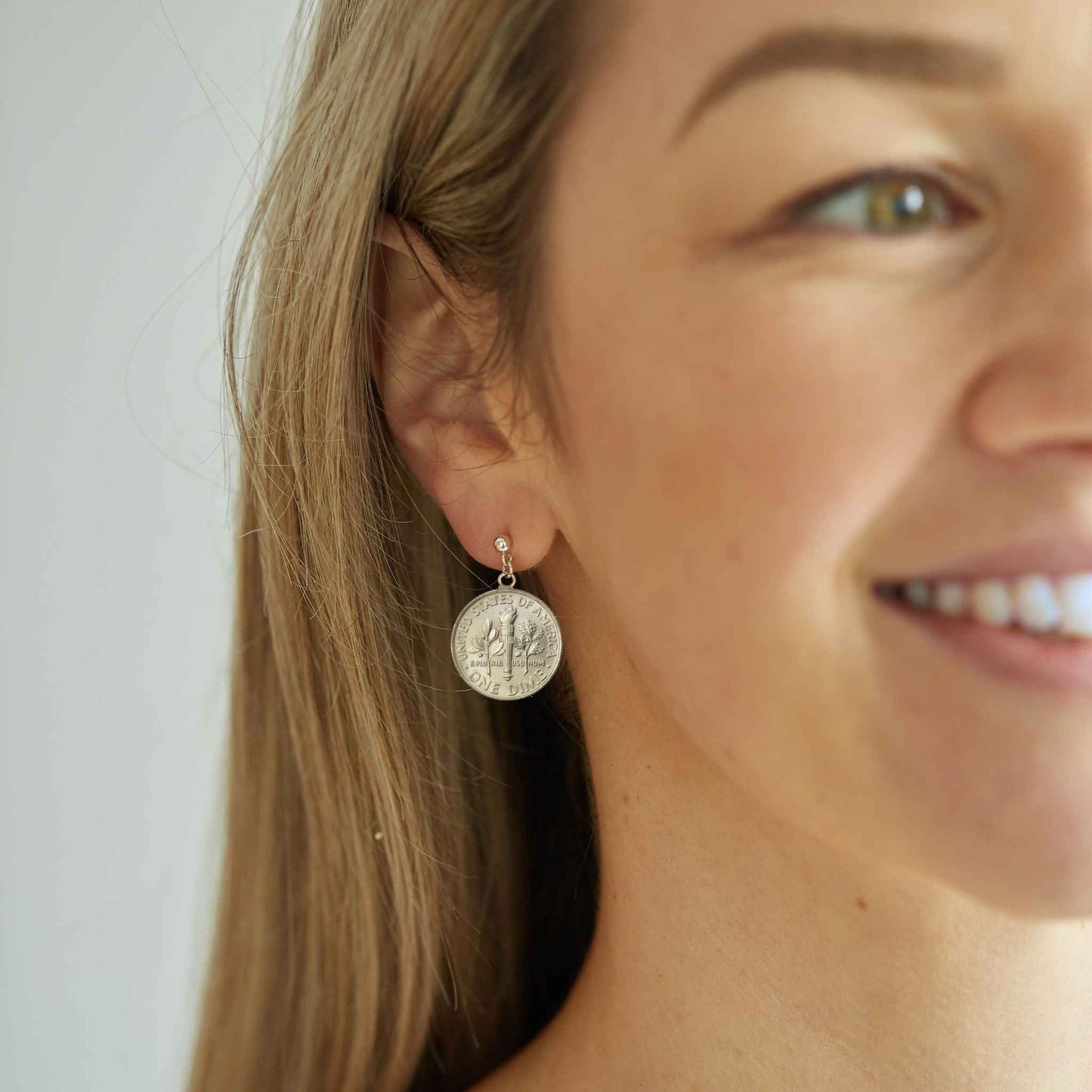 Close-up of a woman wearing silver American Dime coin earrings with a plain background