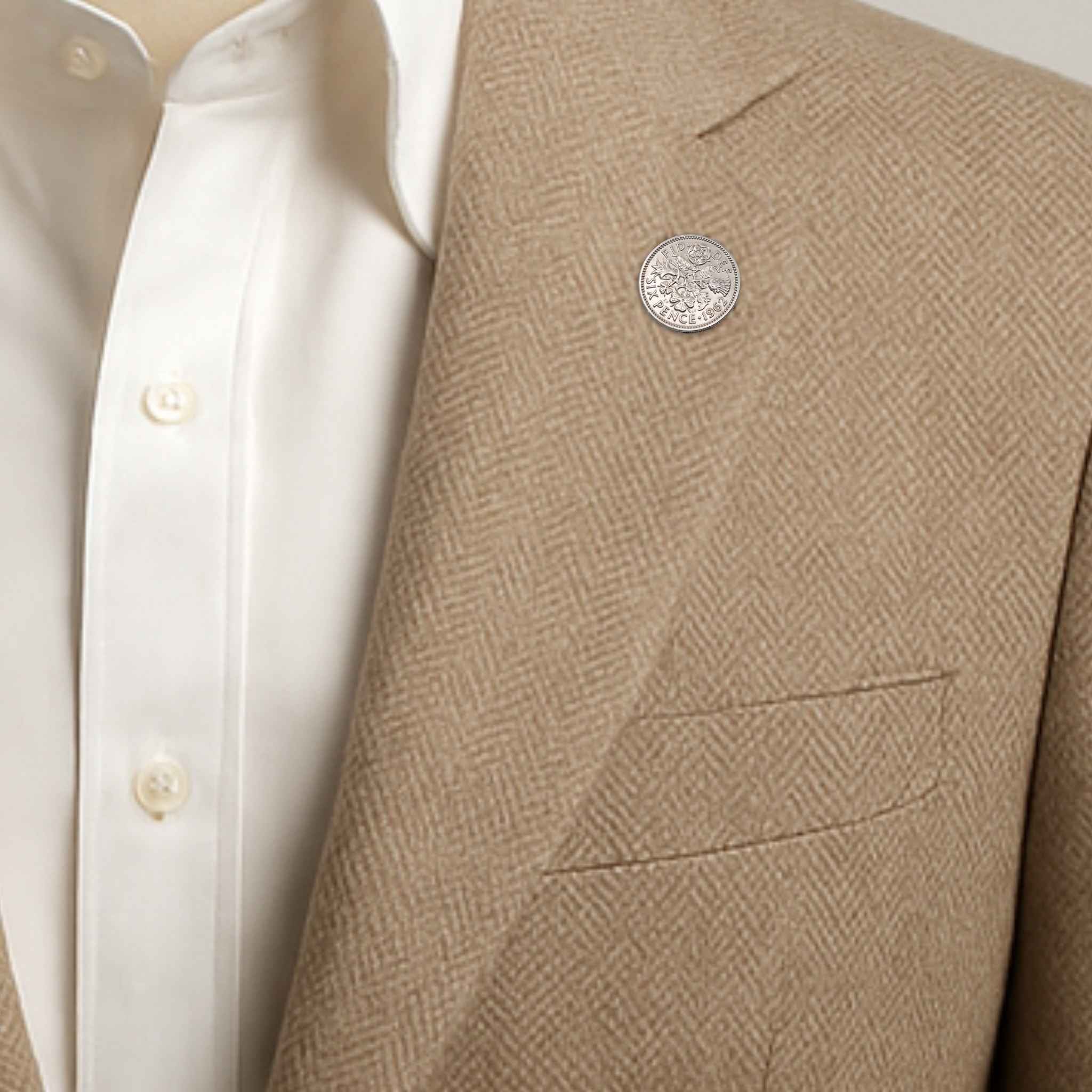 Close-up of a beige blazer with a textured pattern and a Queen Elizabeth 2nd sixpence coin lapel pin button, worn over a white shirt.