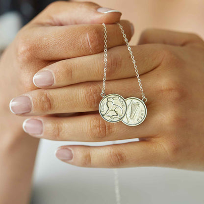 Hand holding a silver necklace with two Irish threepence coins against a neutral background