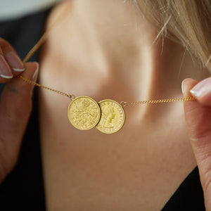 A close-up of a person wearing a necklace featuring two gold-plated sixpence coins on a chain, with one side of the coin showing a floral design and the other side Queen Elizabeth II.